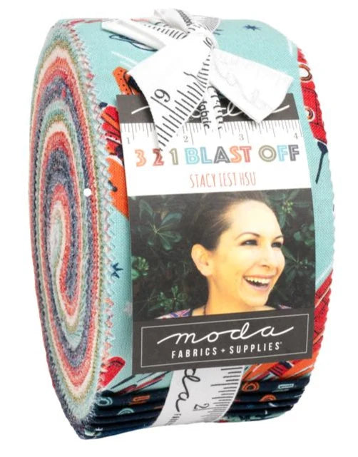 A jelly roll of Moda Fabrics 'Blast Off' fabric by Stacy Lest Hsu, featuring a colorful spiral pattern on the left side and a light blue base with red, orange, and black abstract patterns on the right. The roll is wrapped in a white paper band with a black and white measuring tape graphic and a photo of a smiling woman.
