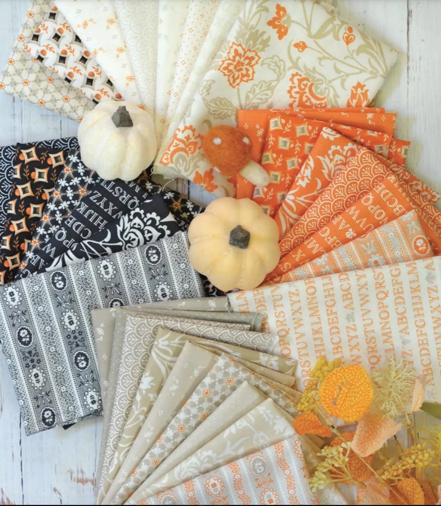 A flat-lay arrangement of multiple fabric designs with autumn-themed patterns in orange, beige, black, and white, styled with two small pumpkins and faux flowers on a white wood background.